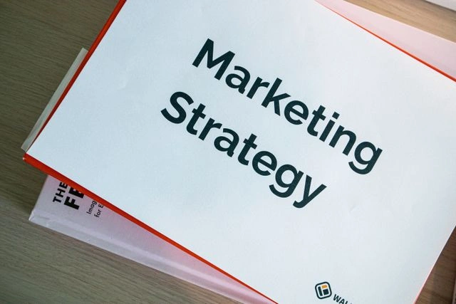 Printed document titled "Marketing Strategy" in bold black text on a desk, resting on top of a soft pink book or binder with an orange border visible.
