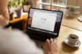 Over-the-shoulder view of a person using a MacBook open to the Google search homepage on a wooden desk next to a coffee cup.