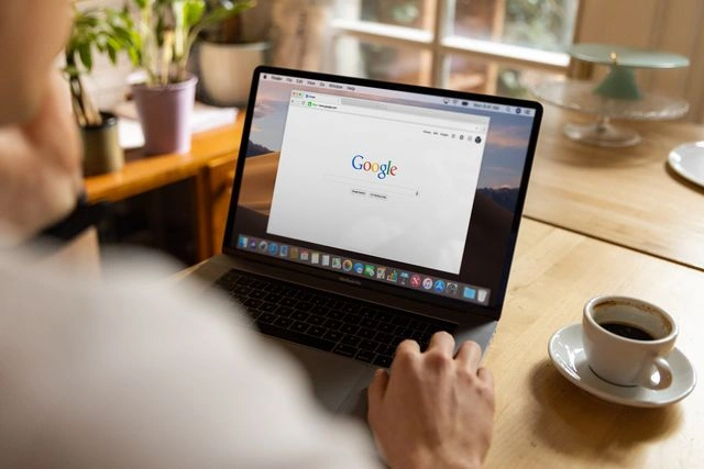 Over-the-shoulder view of a person using a MacBook open to the Google search homepage on a wooden desk next to a coffee cup.