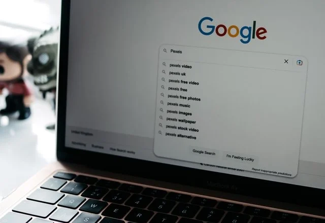 An angled close-up of a open MacBook Air displaying the Google homepage on its screen. The search bar shows "Pexels" typed in, with a dropdown menu displaying search suggestions like "pexels video" and "pexels free photos".
