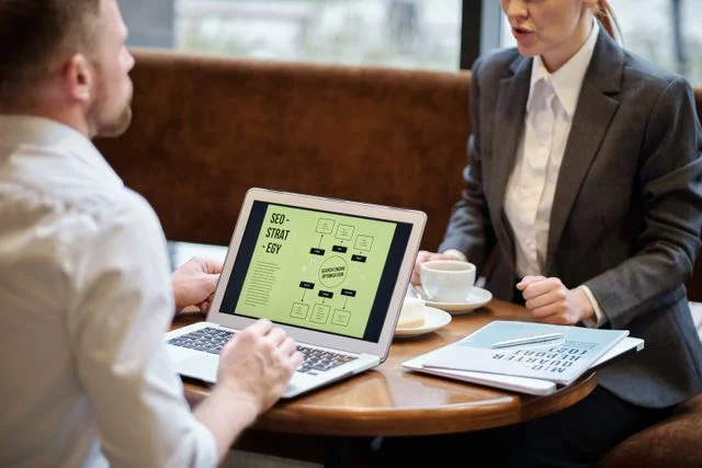 A man and a woman in professional attire sitting at a cafe table discussing a presentation. The man is looking at a laptop showing an "SEO-STRATEGY" flowchart on the screen.