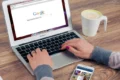 A person typing on an open MacBook Air displaying the Google search home page next to a cup of coffee and a smartphone.