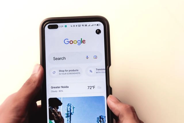 A person holding a smartphone displaying the Google homepage app with a search bar, weather widget showing Greater Noida, and shortcut buttons.