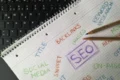 Spiral-bound notebook with handwritten SEO terms like "SEO," "Backlinks," and "Keyword Research" placed next to a black laptop keyboard and colored pencils.