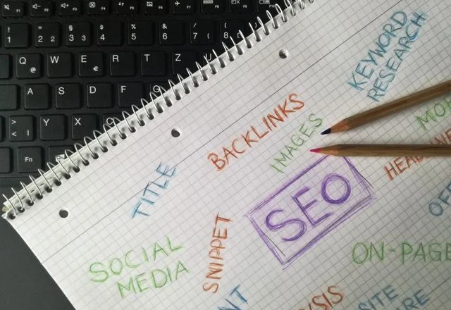 Spiral-bound notebook with handwritten SEO terms like "SEO," "Backlinks," and "Keyword Research" placed next to a black laptop keyboard and colored pencils.