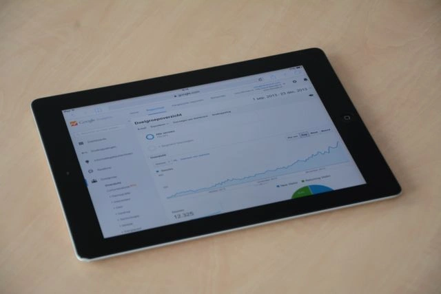 Black iPad resting on a light wooden surface, displaying a Google Analytics dashboard with line graphs and audience metrics in Dutch.